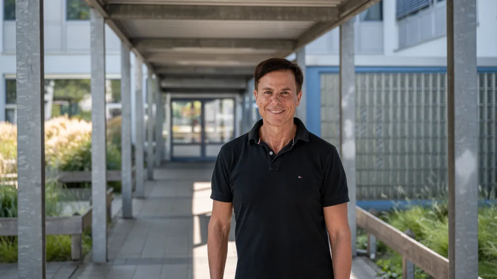Tino Desogus steht im Zentrum eines überdachten Durchgangs vor einem modernen Schulgebäude der Geschwister-Scholl-Schule in Rodgau. Er trägt ein dunkelblaues Poloshirt und lächelt direkt in die Kamera. Der Gang ist von Metallstützen und Glasbaustein-Wänden eingerahmt, auf beiden Seiten wachsen Ziergräser in bepflanzten Holzbeeten. Durch die perspektivische Tiefe wirkt das Bild symmetrisch und klar strukturiert. Die Szene ist hell und freundlich, das Tageslicht betont die Offenheit der Architektur.