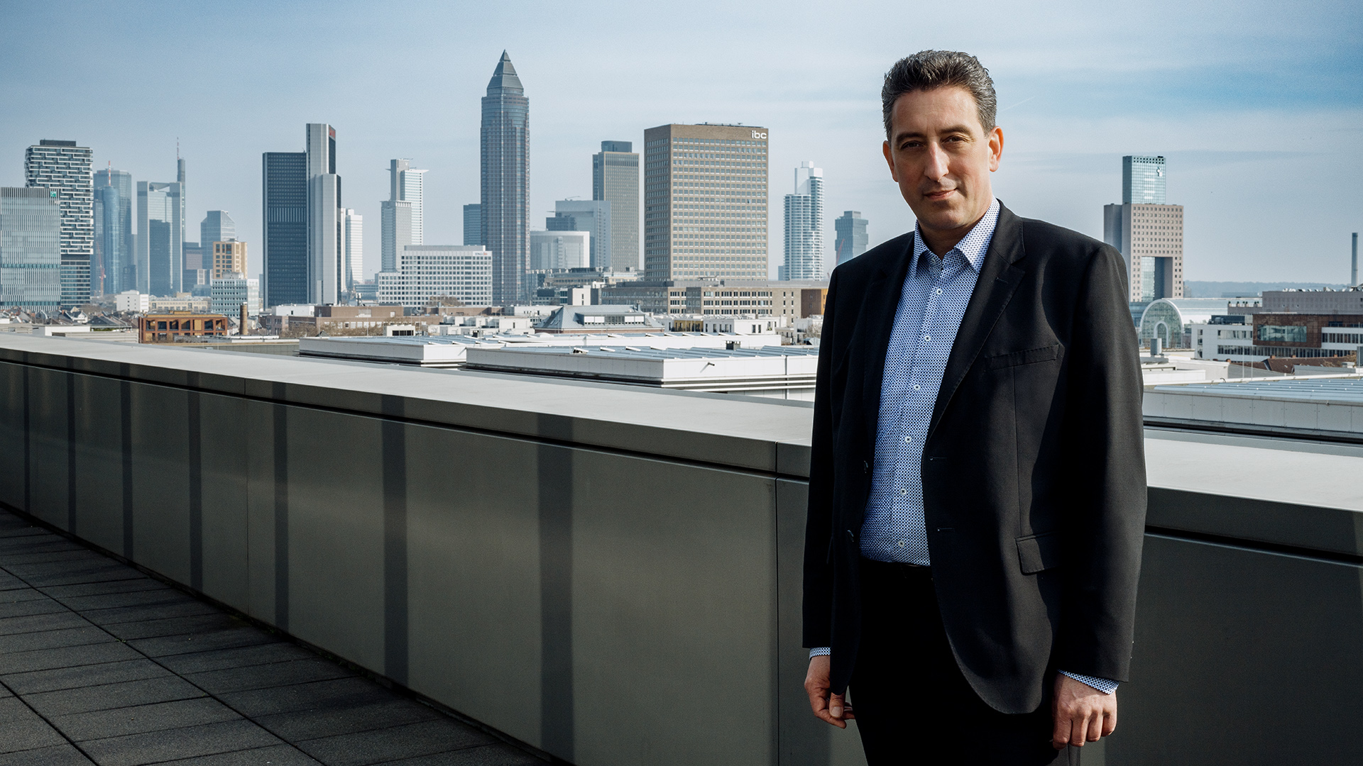Maxelon, Vertreter von Mainova, steht im Business-Outfit auf einem Dach mit Panoramablick auf die moderne Skyline von Frankfurt. Die Aufnahme vermittelt urbanes Flair und symbolisiert wirtschaftliche Stärke sowie regionale Verbundenheit.