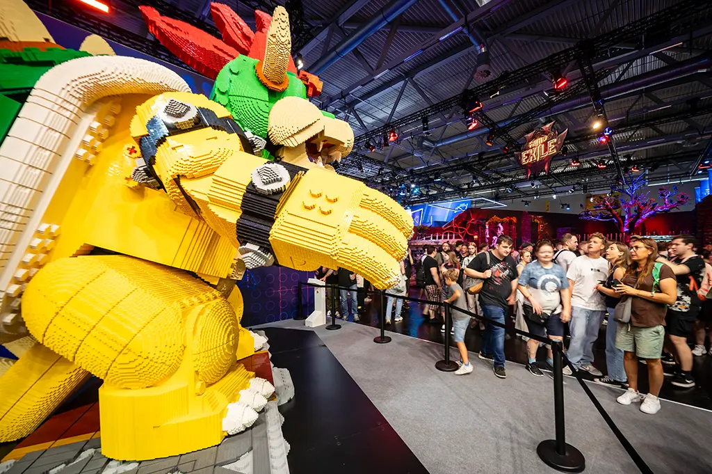 Stand: LEGO GmbH, entertainment area, Halle 6
Das Bild zeigt eine imposante Bowser-Skulptur aus bunten LEGO-Steinen auf der Gamescom. Die Figur ist detailreich gestaltet, in leuchtendem Gelb, Grün, Rot und Schwarz, und ragt eindrucksvoll in die Messehalle. Auf der rechten Bildseite beobachten zahlreiche Besucherinnen und Besucher die Figur aus nächster Nähe, während im Hintergrund bunte Beleuchtung und weitere Messestände zu sehen sind. Die Szene vermittelt die kreative und verspielte Atmosphäre des Gaming-Events.