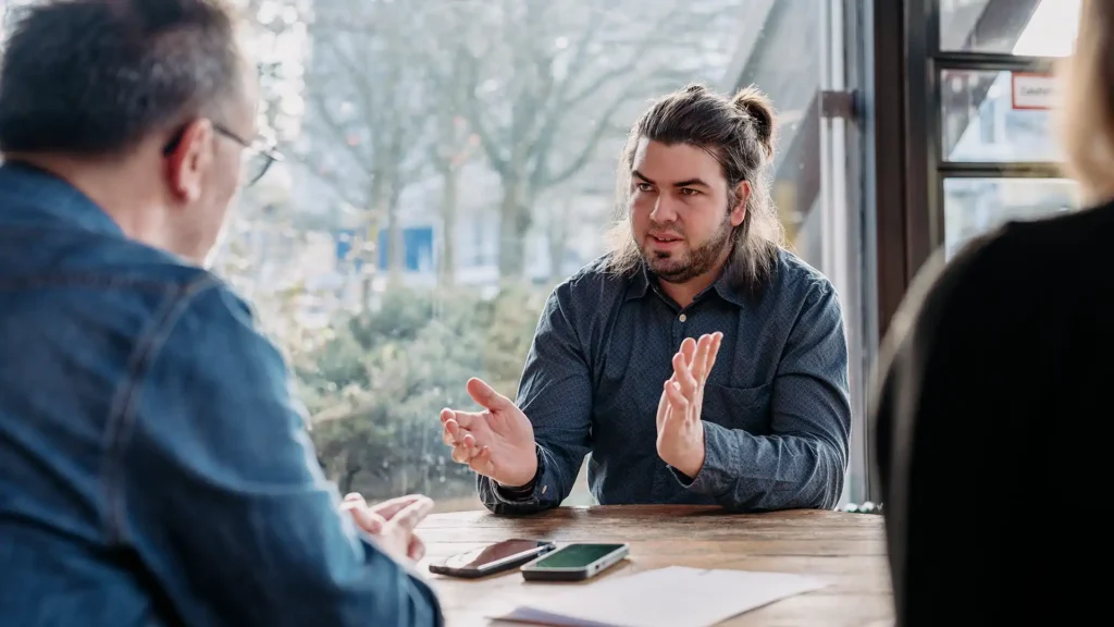 Personen im Gespräch an einem Holztisch vor einer großen Fensterfront. Alexander gestikuliert, während er seine Idee erklärt.