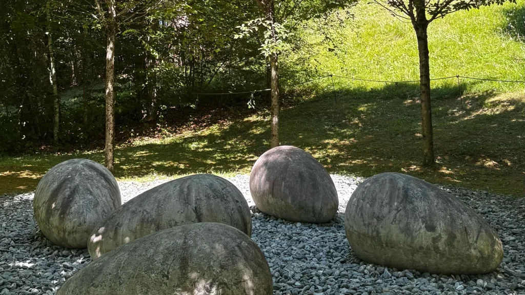 Große steinerne Eier als Kunstinstallation im Freilichtmuseum Arte Sella in Trentino, umgeben von Bäumen und Kies.