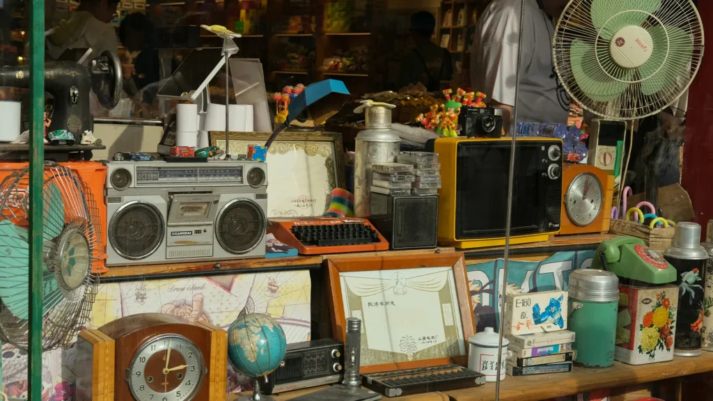Ein vollgestellter Stand mit einer Sammlung von Vintage-Gegenständen, darunter ein großer, silberner Ghettoblaster, ein kleiner gelber Röhrenfernseher, alte Tischuhren, eine orangefarbene Schreibmaschine, Ventilatoren und ein kleiner Globus – diese schönen Alltagsgegenstände erzählen Geschichten aus der Vergangenheit.
