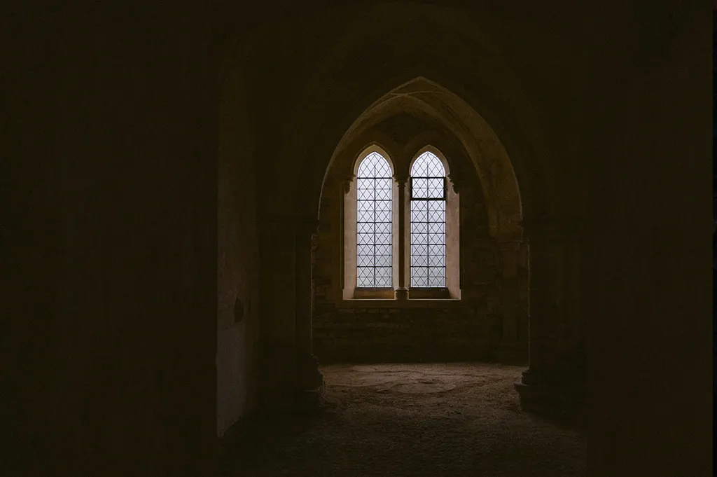 Schwach beleuchteter Raum mit gotischem Spitzbogenfenster und alten Steinmauern. Die geheimnisvolle Stimmung passt zur mittelalterlich inspirierten Musik des Dungeon Synth.